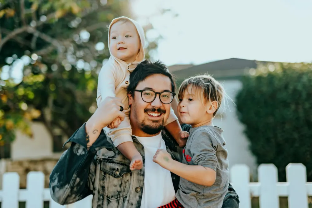 A smiling father outdoors carrying his two young children by a white picket fence